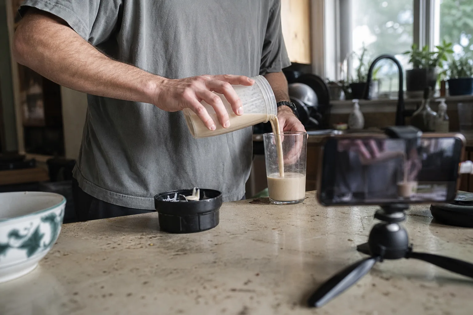 Hands pouring a protein shake while a phone on a mini tripod films it